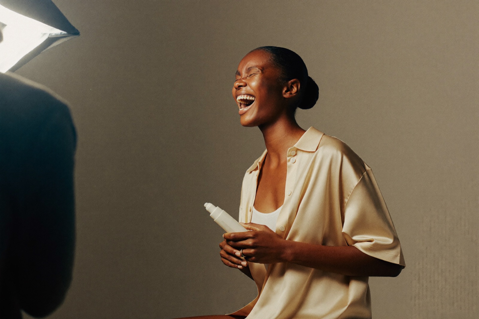 A model shooting a lifestyle e-commerce campaign against a plain studio backdrop, mid-laugh, product in hand