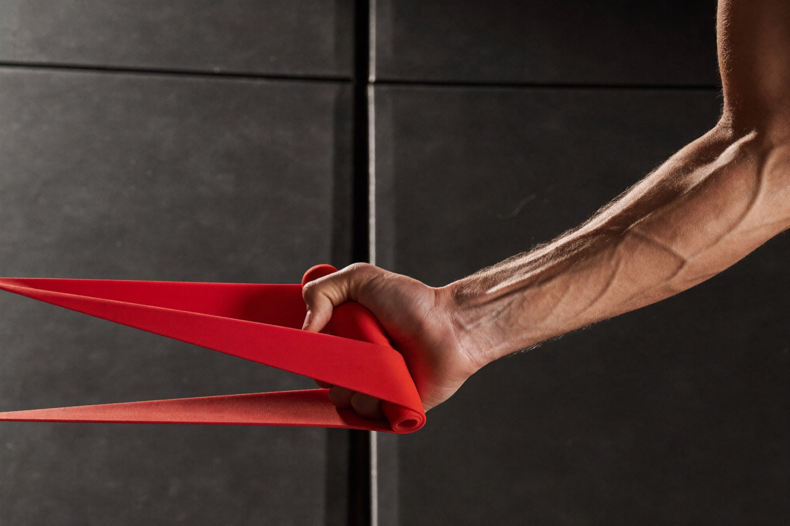 A close crop of a model's hand gripping a resistance band mid-exercise, gym floor visible in soft focus behind