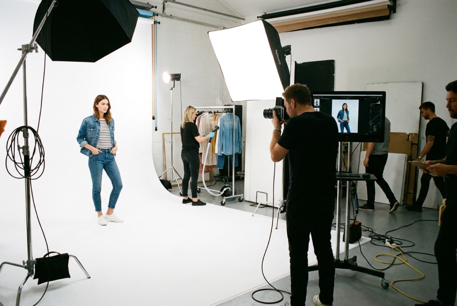 A commercial e-commerce studio mid-shoot with a model on a white backdrop and photographer at a tethered monitor