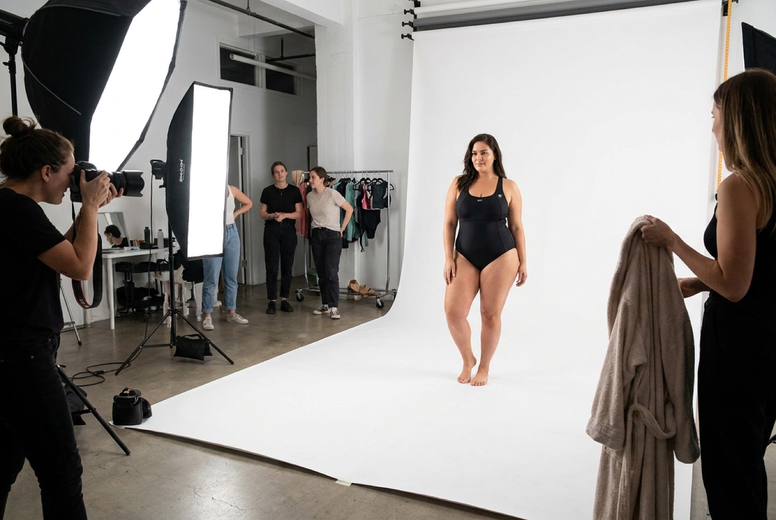 A swimwear e-commerce shoot in a professional studio with a plus-size model on a white cove, stylist holding a robe between frames